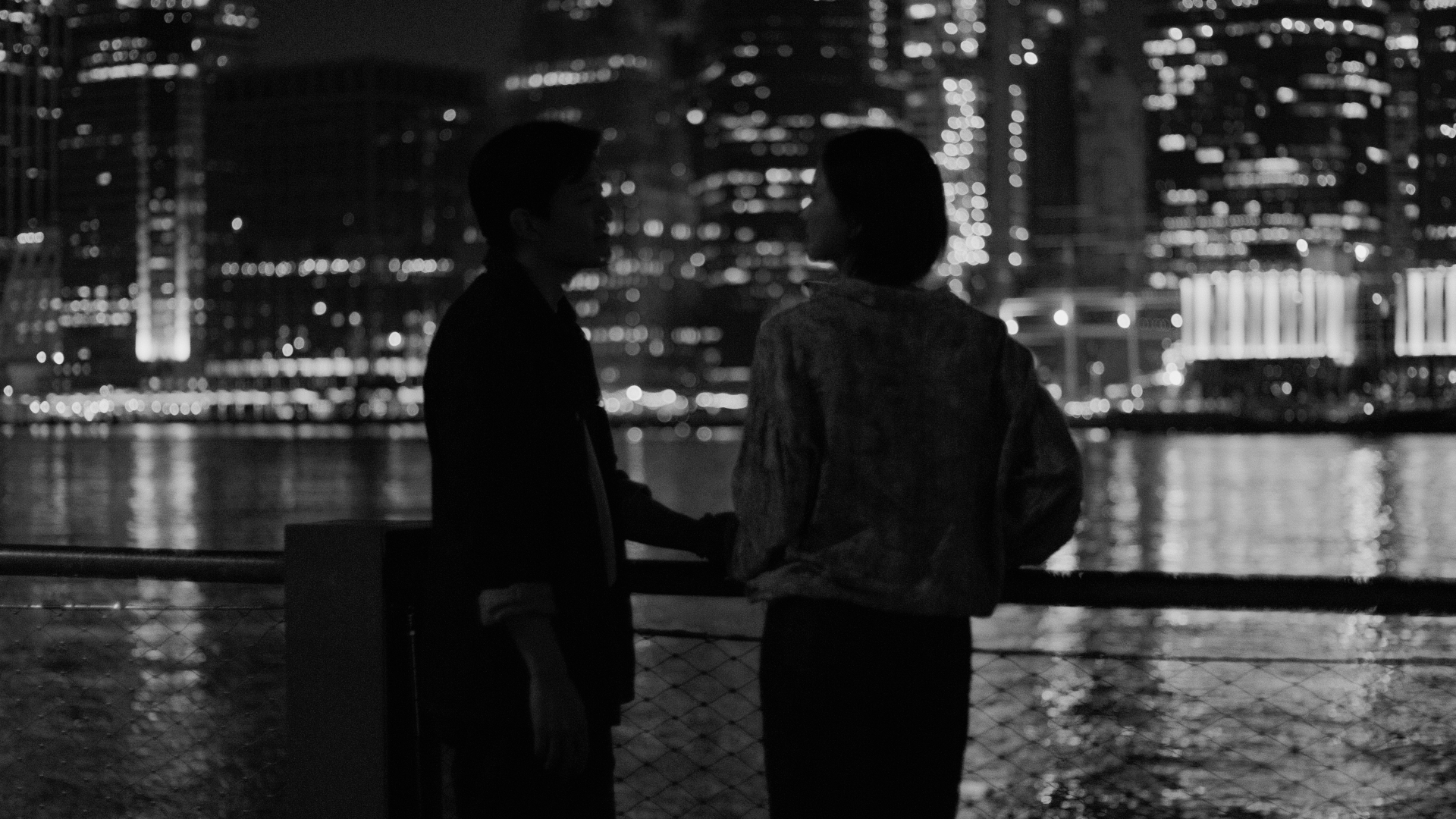Two figures by the water, city lights behind them at night